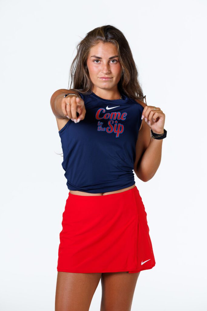 A person in athletic attire points forward while wearing a navy blue top with "Come to the Sip" printed on it and a red skirt.